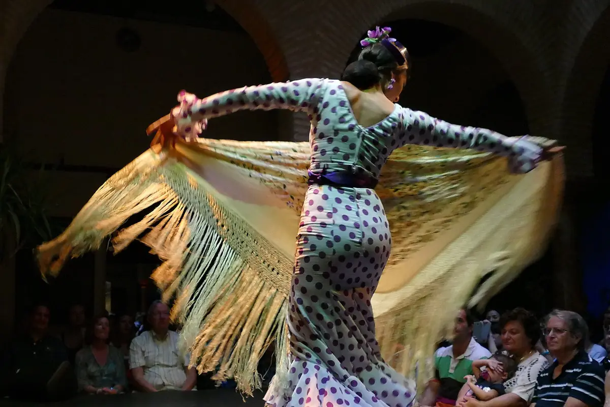 Eine Flamenco-Tänzerin im gepunkteten Kleid wirbelt einen langen Fransenshawl über eine dunkle Bühne, während das Publikum ganz nah dabei ist