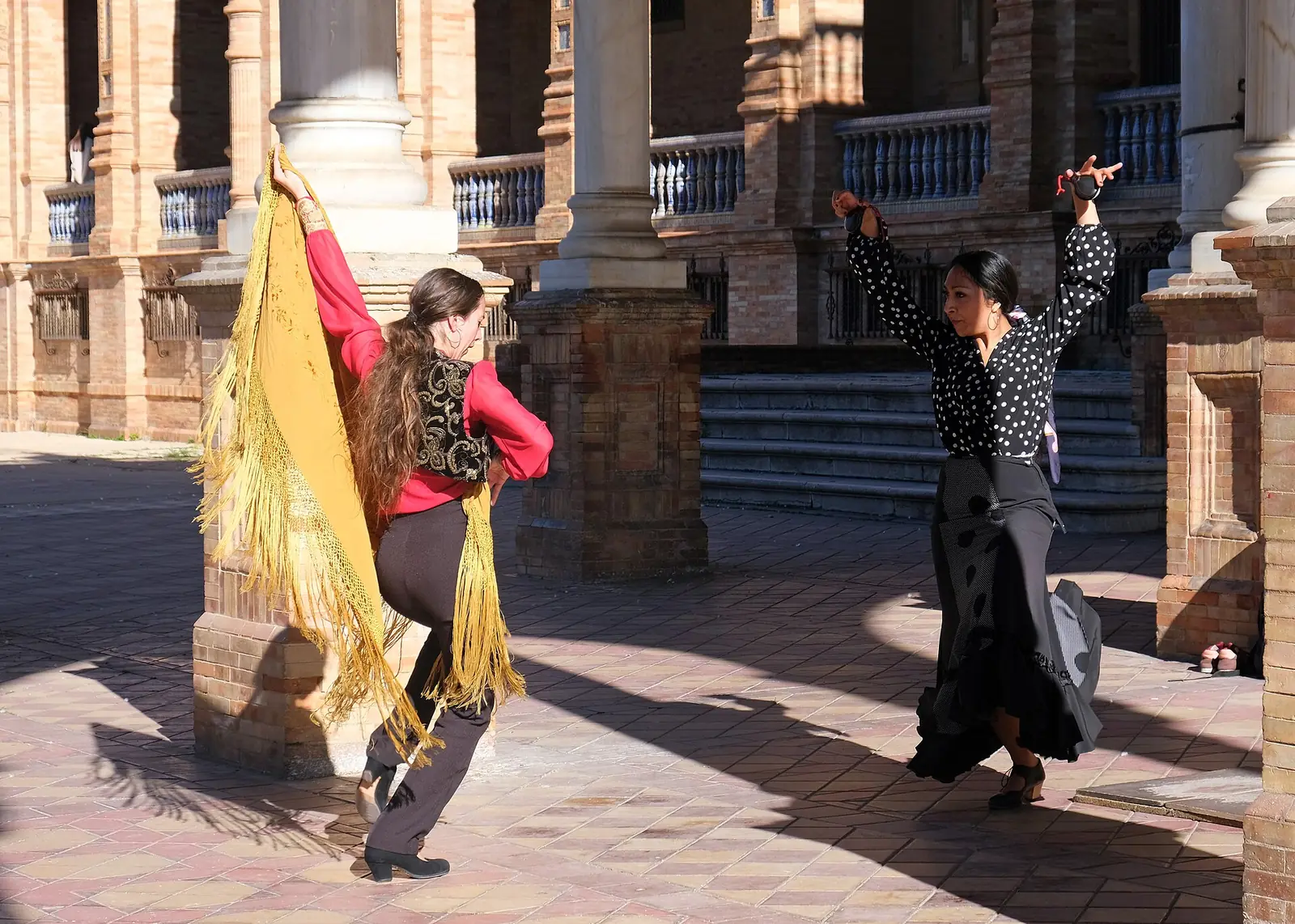 Dva flamenkoví tanečníci, jeden se zlatým šálem a jeden s kastanětami, tančí v cihlové arkáde na Plaza de España v Seville